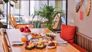 Una mesa de comedor dispuesta con alimentos para el desayuno, como frutas, pan y bebidas, en una habitación con un ventanal grande y una planta.