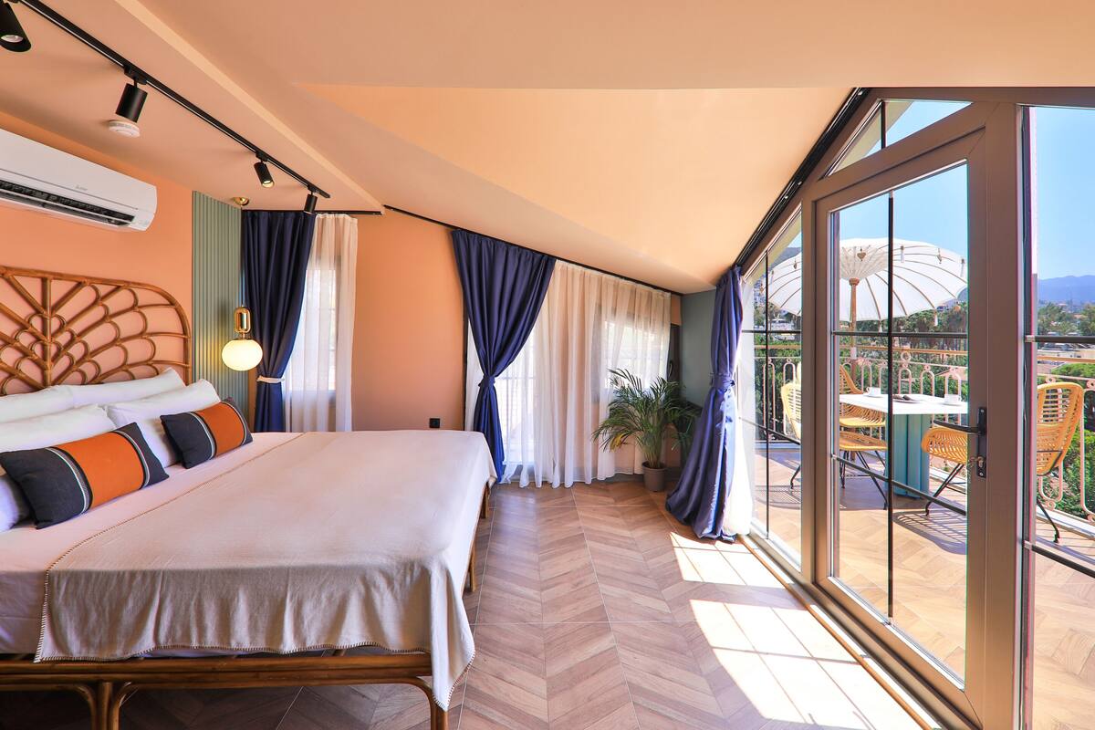 Habitación de hotel con una cama grande, un balcón con vista a un parque y una zona de comedor con muebles de exterior.