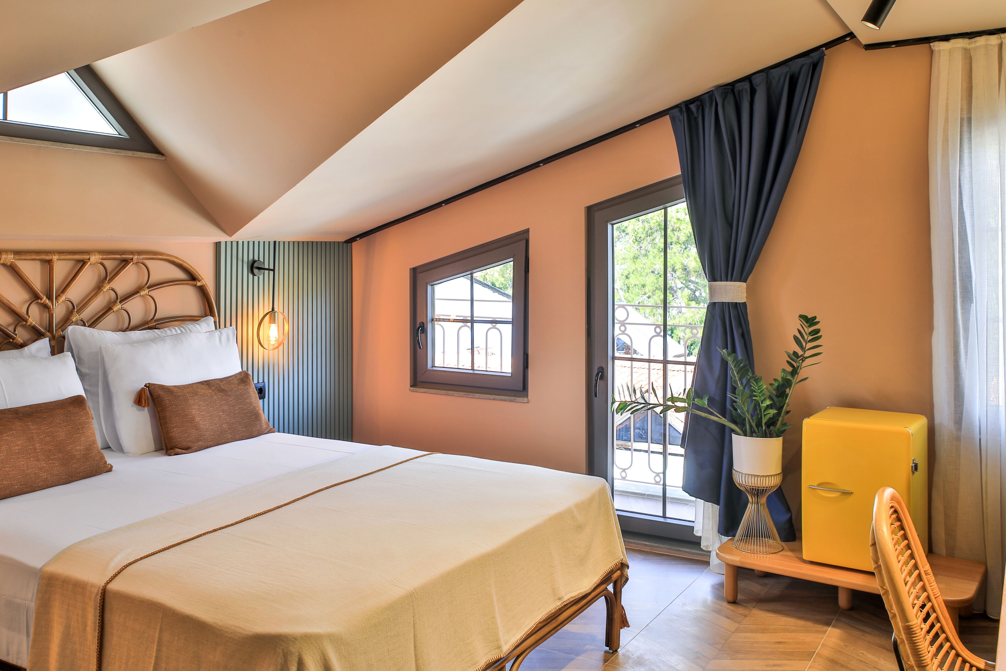 Une chambre avec un grand lit, une petite armoire jaune, une chaise en osier et une plante en pot.