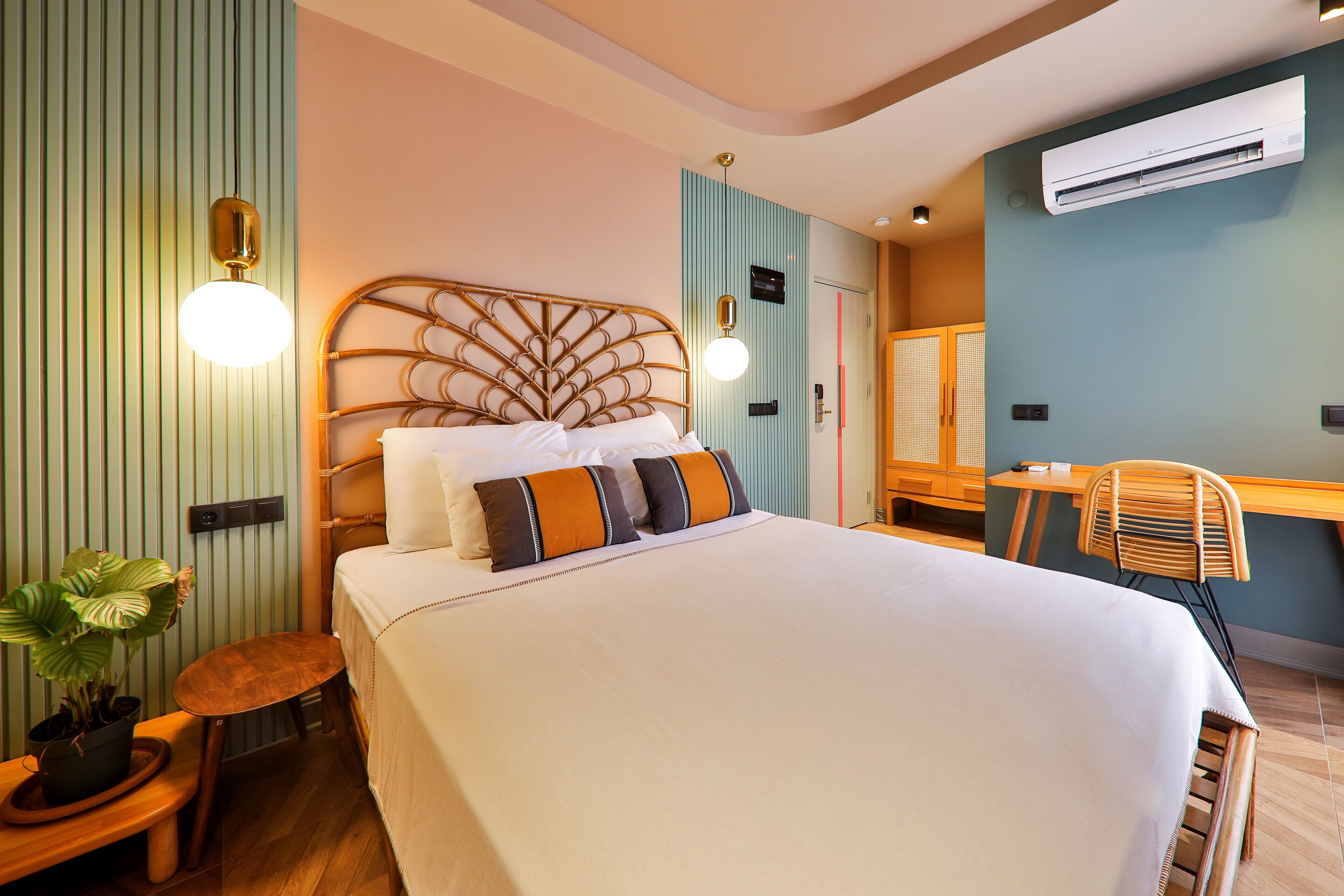 A hotel room with a bed, bedside table, potted plant, and a desk with a chair.