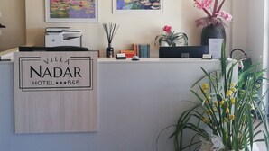 A hotel reception desk with framed paintings, a sign reading "Villa Nadar Hotel B&B", and various decorative items including plants and books. - Villa Nadar Hotel  B&B (Altopascio)