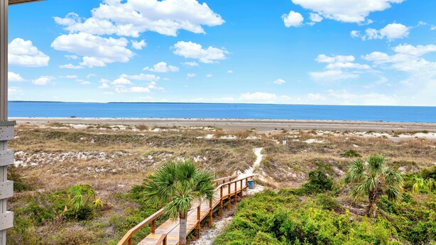 Eine Holzterrasse, die zu einem Strand f ührt, mit freiem Blick auf das Meer und einem Sandweg.