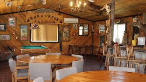 Ein rustikaler Innenraum mit Holzwänden und -decke, einem Billardtisch sowie mehreren Esstischen und Stühlen.
