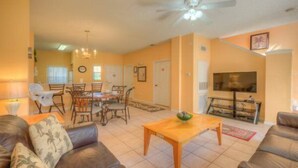 A living room with a sofa, a coffee table, a dining area with chairs, a ceiling fan, and a television. - Emerald_island_8515n (Kissimmee)