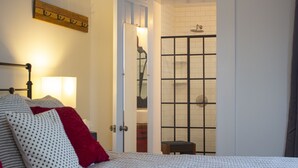 A bedroom with a bed, a nightstand, a lamp, a red pillow, and a door leading to a bathroom with a shower.