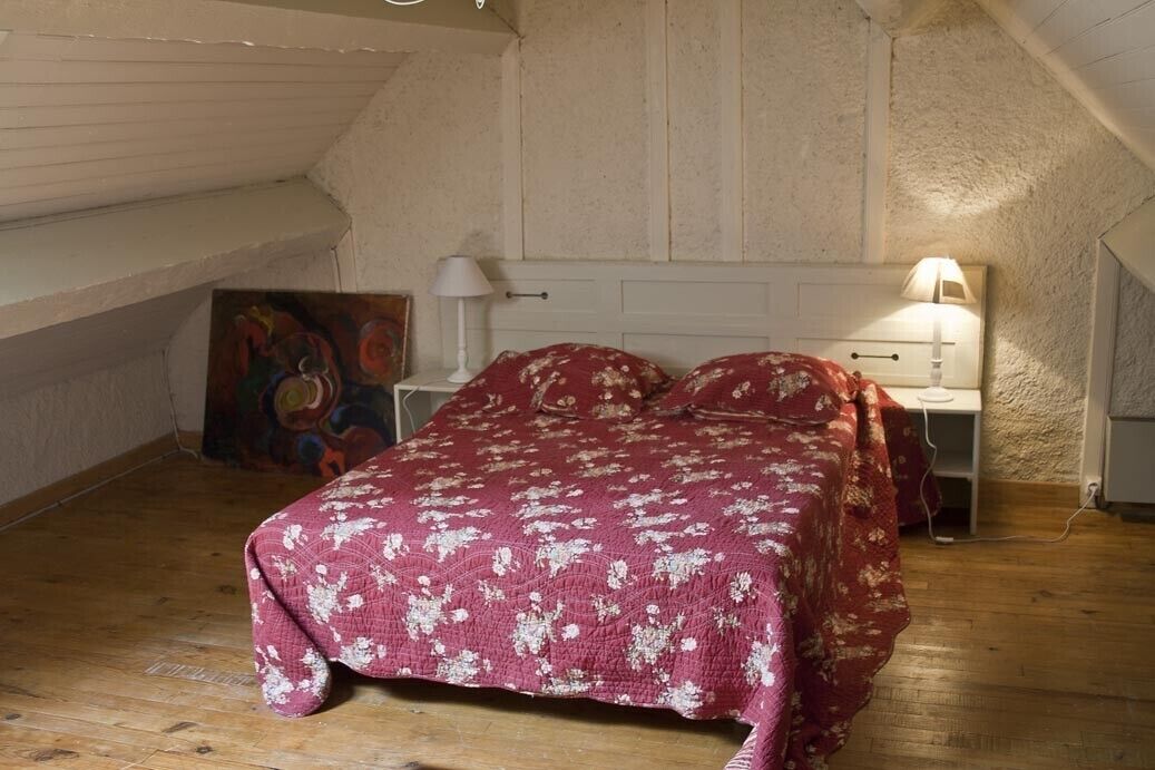 A bedroom with a wooden floor, a bed with a red floral bedspread, a nightstand with a lamp, and a painting on the wall.