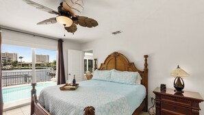 Un dormitorio con una cama grande, una mesita de noche, una lámpara y vistas a una piscina y edificios.