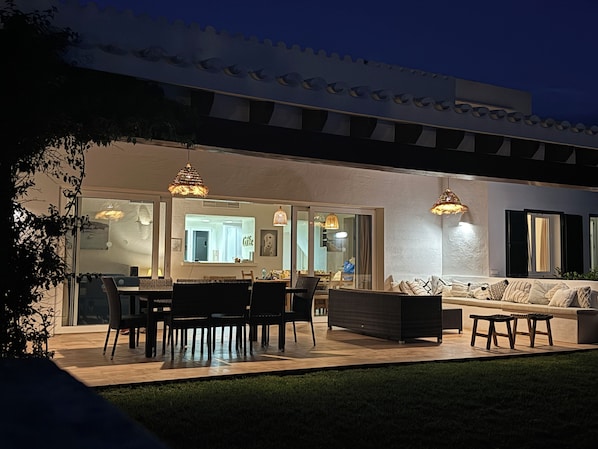 An outdoor patio area with a dining table, chairs, and a sofa. There are hanging lights and a view into the house. - Binibeca - Luxurious Villa Esmee - Swimming Pool -Wifi - 10 persons (Sant Lluís)