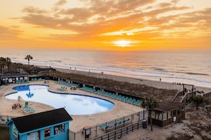 Zona de piscina frente al mar con sillas de descanso, una torre de socorristas y vistas al océano al atardecer.