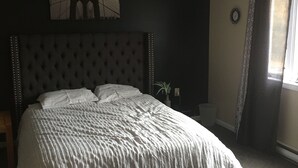 A bedroom with a large bed, a headboard, a wall clock, and a window with curtains.