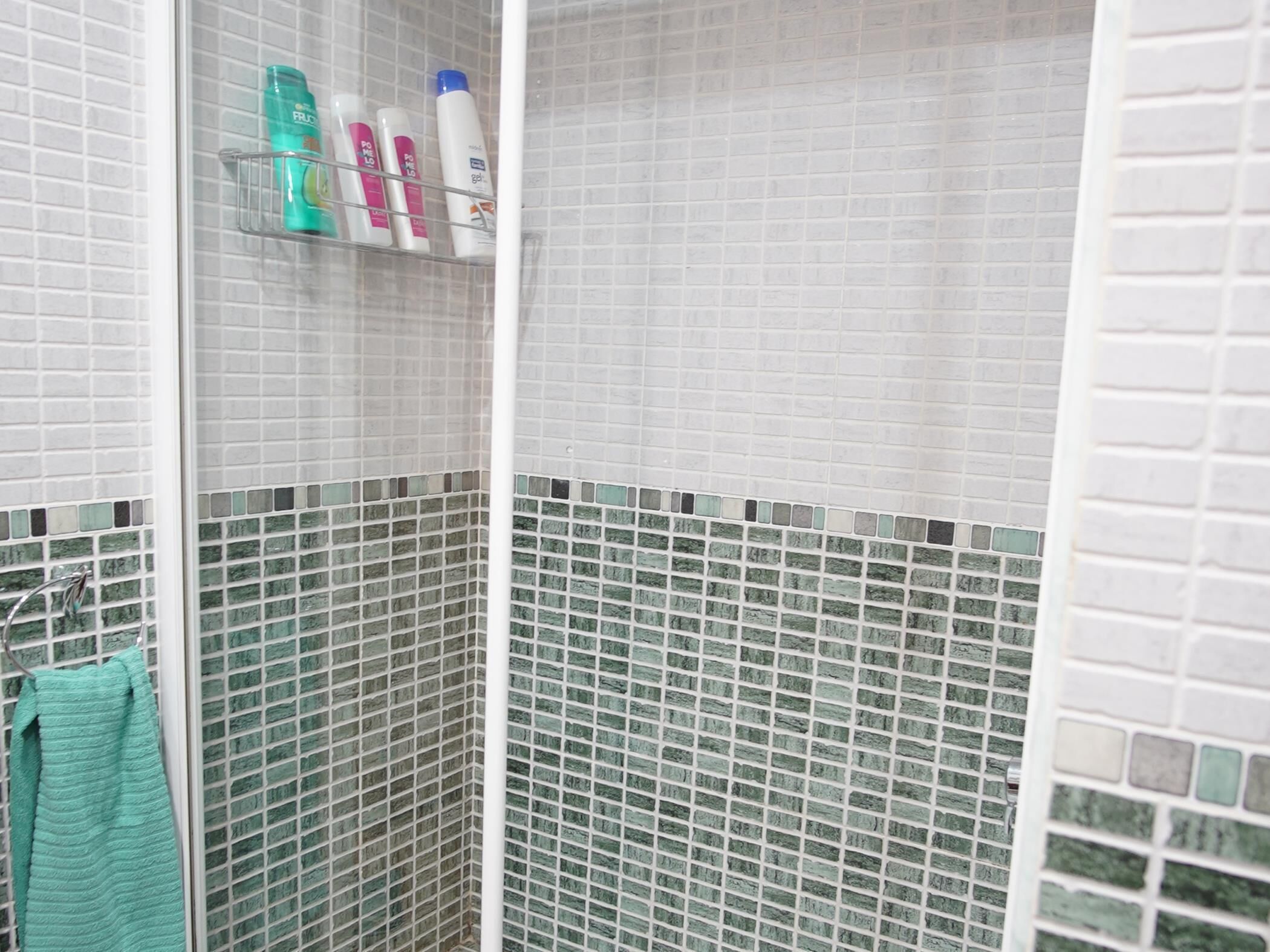 Un baño con paredes de azulejos blancos, una zona de ducha con puerta de cristal y un estante con artículos de aseo.