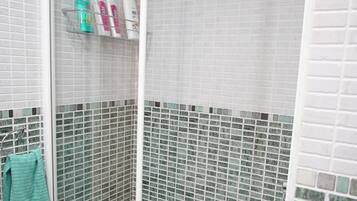 Un baño con paredes de azulejos blancos, una zona de ducha con puerta de cristal y un estante con artículos de aseo.