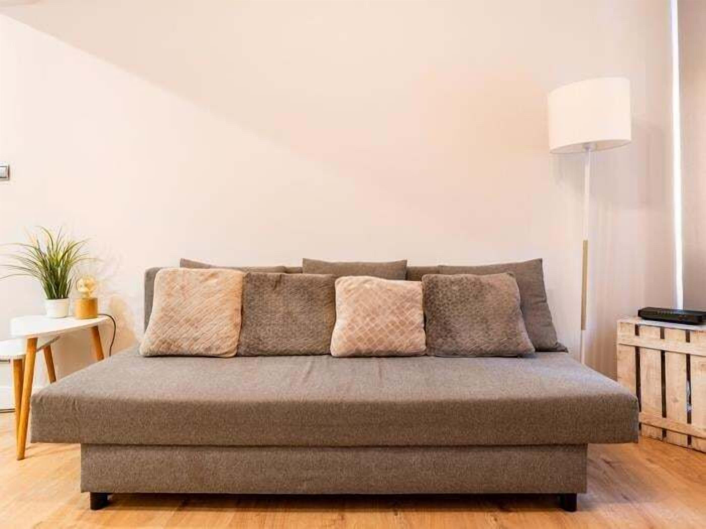 Un sofá cama de tono neutro con un reposapiés a juego, una lámpara de pie y una mesita con una planta.