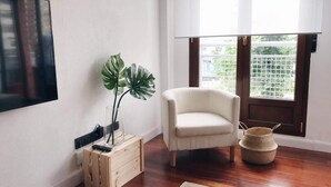 Una habitació amb una armchair blanca, una caixa de fusta amb una planta, una cistella teixida i un televisor fixat a la paret.