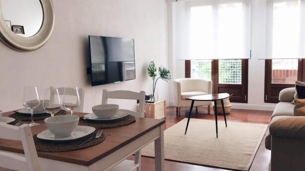 Un saló modern amb zona de menjador, televisió plana penjada a la paret, sofà i finestra gran amb cortines.