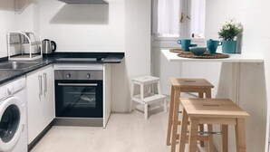 Una cuina funcional amb forn integrat, rentadora i zona de dinar petita amb taula i cadires de fusta.