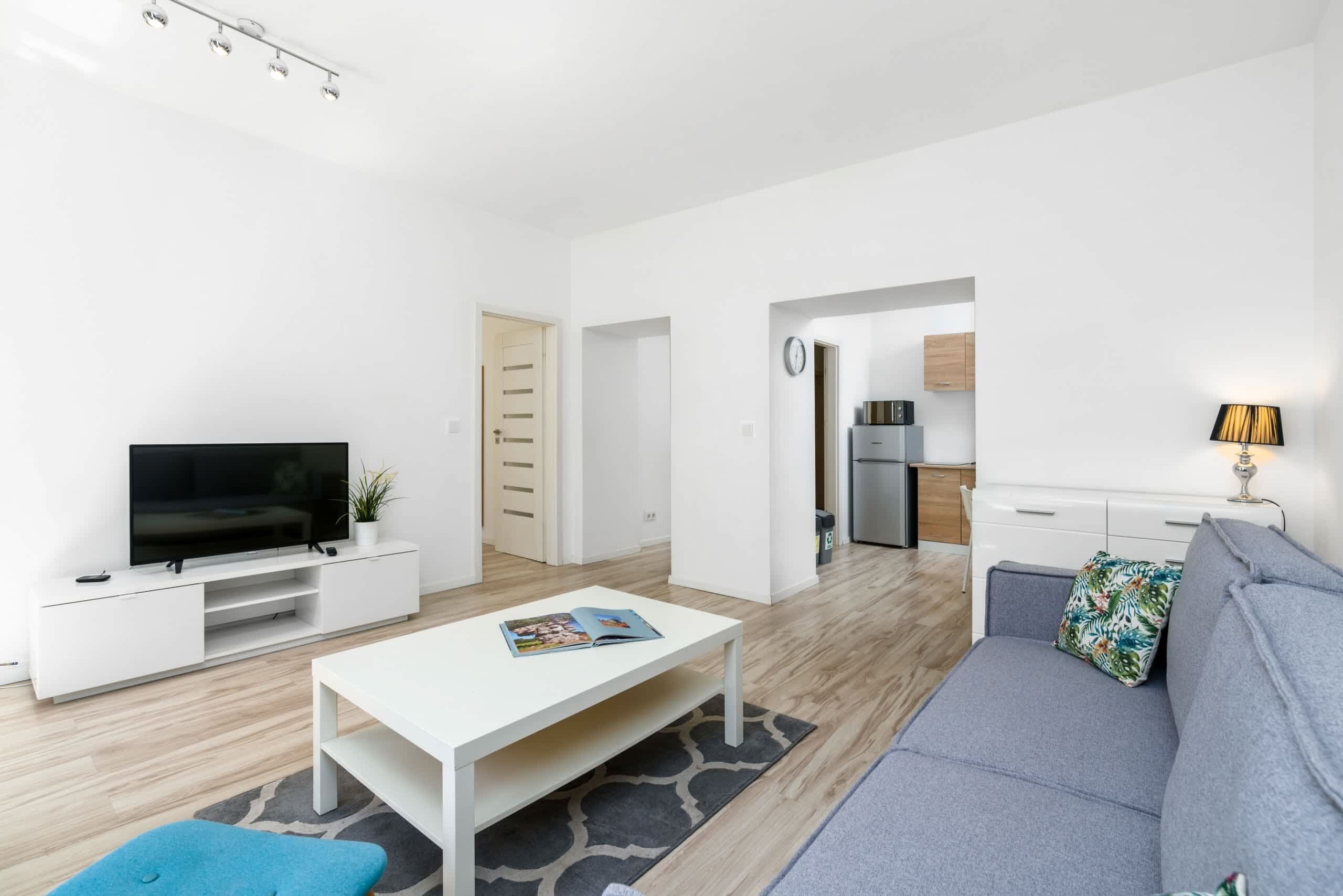 A modern living room with a flat-screen TV, a white coffee table, a grey sofa, and a small blue ottoman. There is a kitchen area visible in the background with wooden cabinets and a refrigerator.