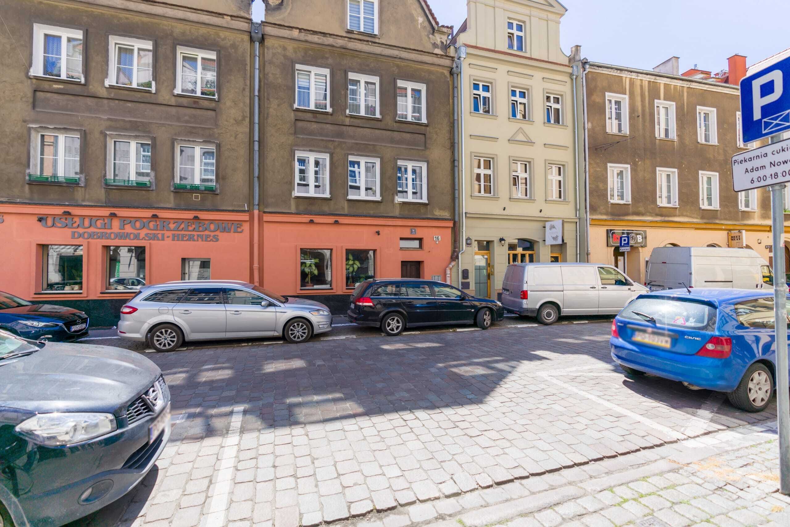 A street view with parked cars, a building with a sign reading "USEGUI POGRZEBOWE DOBROWOLSKI-HIERNES", and a blue parking sign.