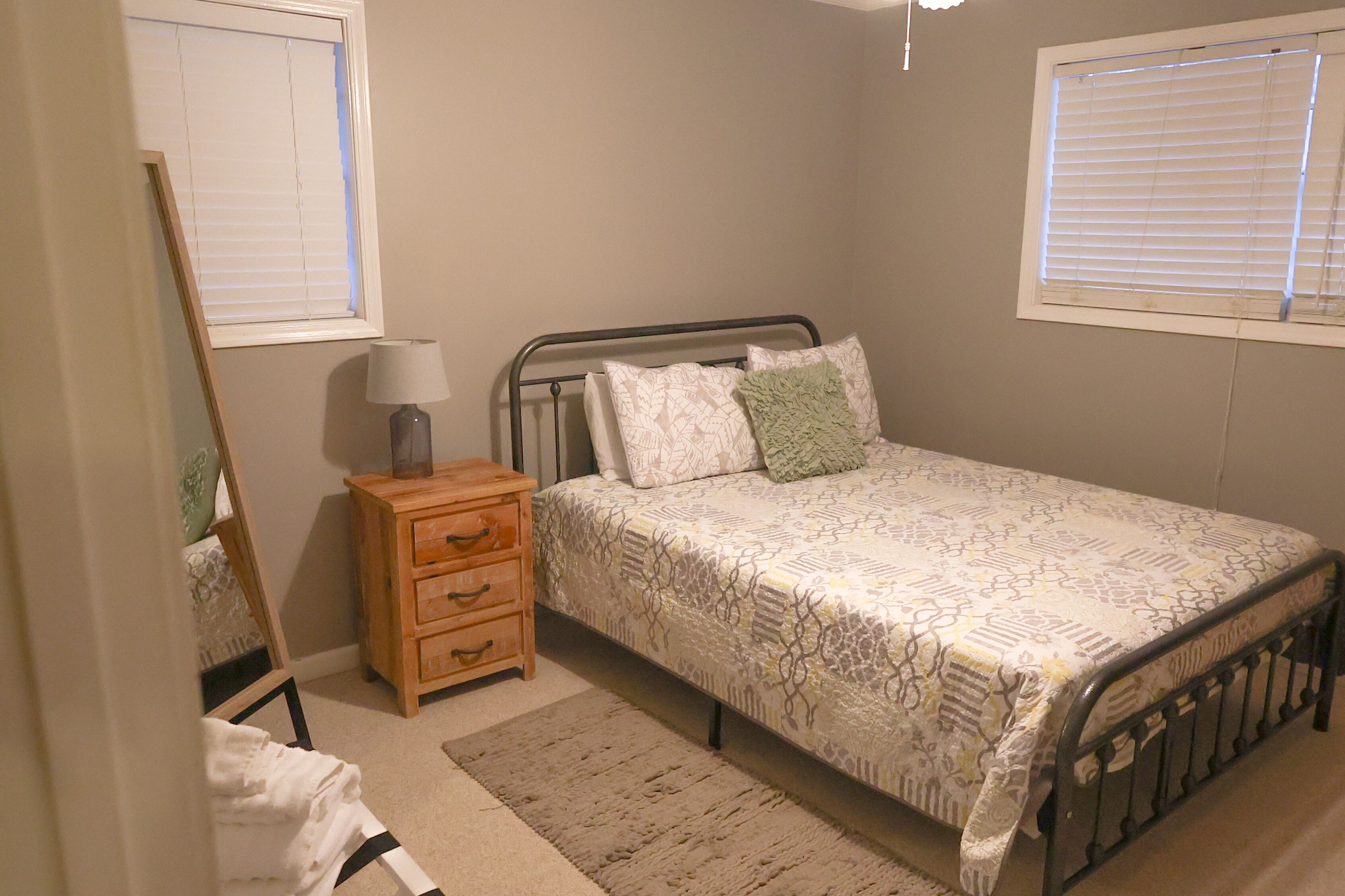 Un dormitorio con cama, una mesita de noche de madera con lámpara, un espejo y una ventana con persianas.