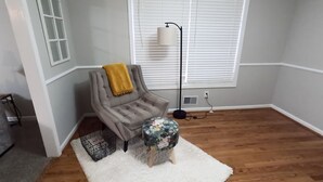 Una habitación con un sillón gris, un reposapiés pequeño y redondo, un tapete blanco y mullido, una lámpara de pie y una ventana con persianas.