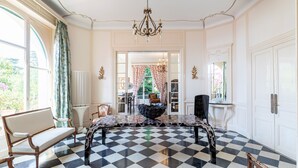 Ein Zimmer mit Schachbrettmuster-Boden, einer Kristallleuchter, einem großen Tisch und Blick auf einen Garten durch ein Fenster.