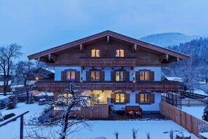A two-story wooden house with a balcony, snow-covered surroundings, and a mountainous backdrop.