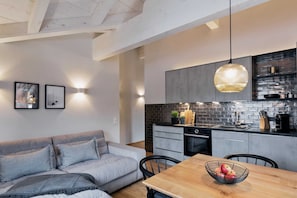 A modern kitchen with a black brick backsplash, a wooden dining table with a fruit bowl, and a gray sofa in a living area.