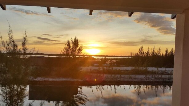 Tramonto su un'acqua calma con alberi riflessi nella superficie, vista da un balcone coperto.