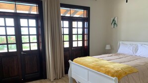 Una camera da letto con pavimento in legno, un letto bianco con coperta gialla, una testiera bianca, una finestra con tende e una lampada a parete.