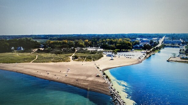 Vue aérienne d’une zone côtière comprenant une plage, un port de plaisance et un quartier résidentiel avec des maisons et des arbres.