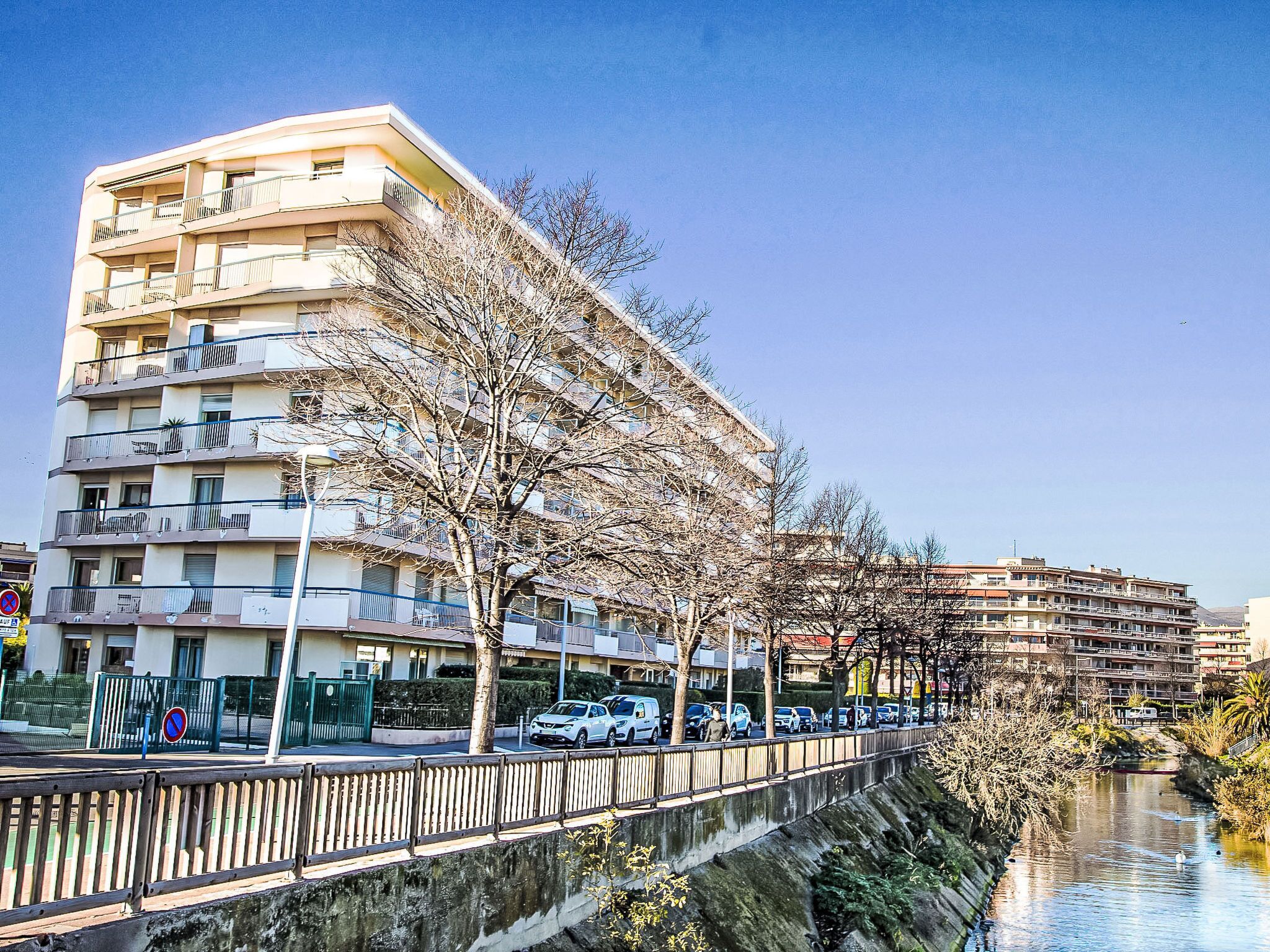 A multi-story building with balconies, a river in the foreground, and parked cars along the street.