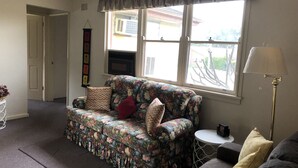 Ein Wohnzimmer mit floraler Couch, einem kleinen runden Tisch, einer Lampe und einem Fenster mit Ausblick.