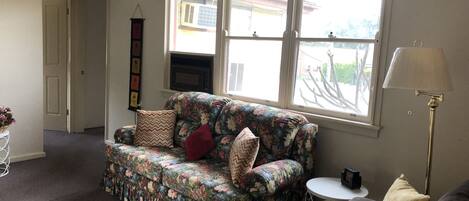 A living room with a floral sofa, a small round table, a lamp, and a window with a view of the outside.