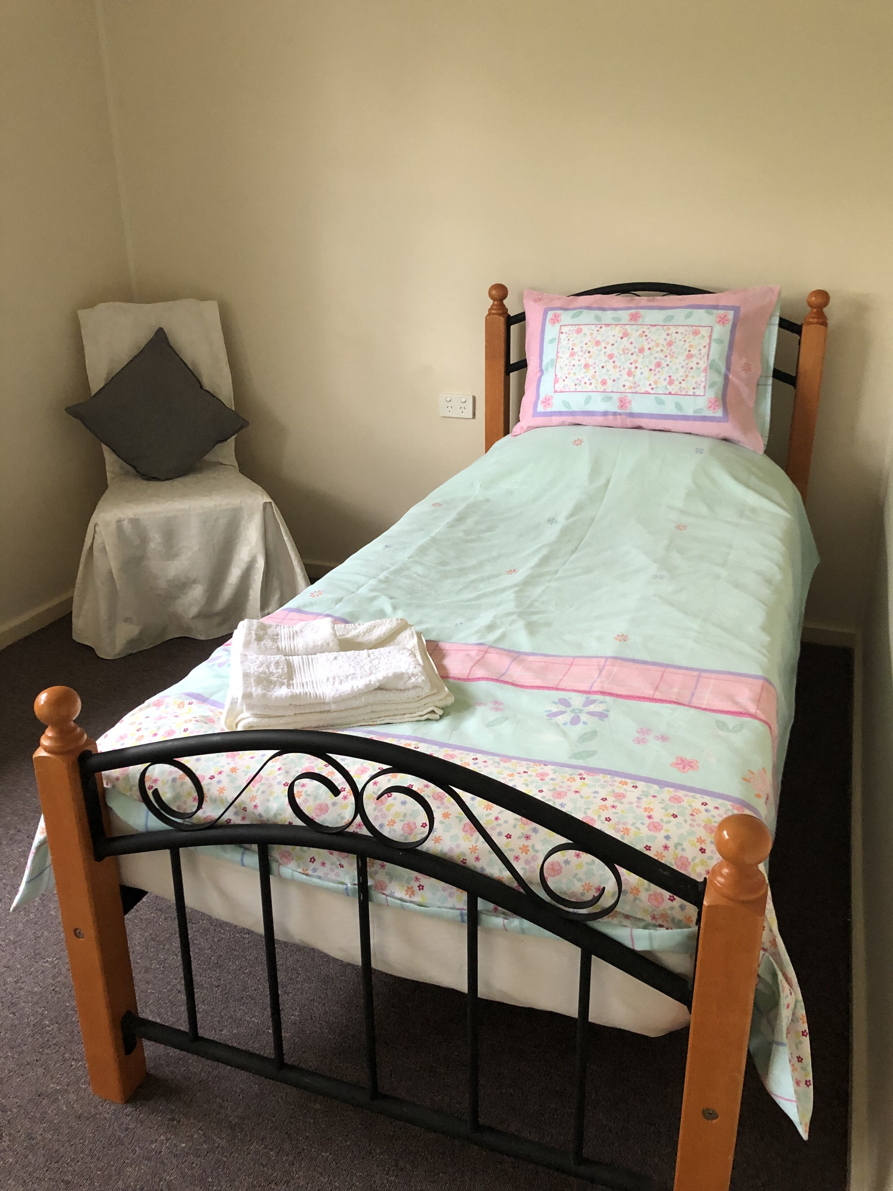 A single bed with a floral bedspread, a wooden headboard, and a small chair with a cushion.