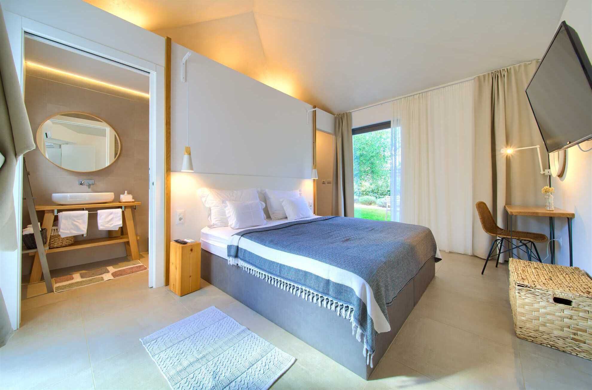 A modern bedroom with a large bed, a bedside table, a mirror, a desk with a chair, and a view of greenery outside.