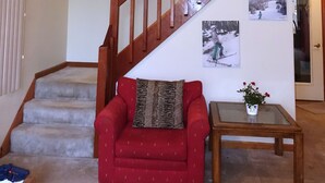 Een woonkamer met een houten trap, een rode bank, een klein tafeltje met een plant en fotokaders aan de muur.