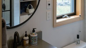 A bathroom with a round mirror, a sink, a window with a view of trees, and a shelf with towels and a basket. - Mountain Condo w Modern CabinStyle @Hunter&Windham (East Jewett)