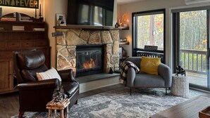 A cozy living room with a stone fireplace, leather chairs, a grey sofa, and a large TV. - Mountain Condo w Modern CabinStyle @Hunter&Windham (East Jewett)