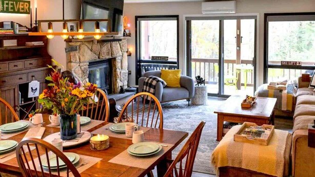 A cozy living room with a fireplace, a dining area with a table set for four, and a sofa with a view of the outdoors. - Mountain Condo w Modern CabinStyle @Hunter&Windham (East Jewett)