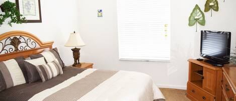 A bedroom with a bed, a nightstand, a lamp, a TV on a wooden stand, a window with blinds, and wall decorations.