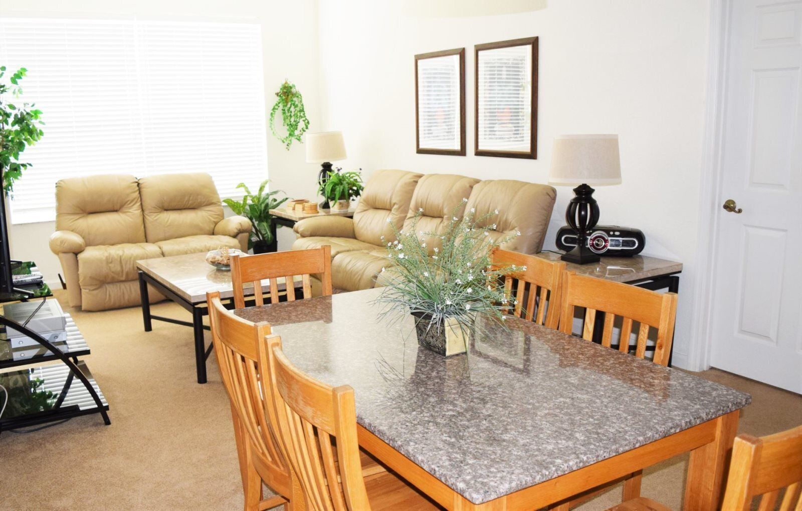 A living room with a beige sofa, a wooden dining table, and a coffee table. There are two lamps, a potted plant, and a framed picture on the wall.