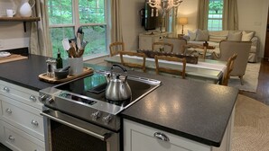 A modern kitchen with a stainless steel stove, a dining area with wooden chairs, a chandelier, and a view of the outdoors through large windows. - Renovated Cottage - Walk to Downtown Mentone (Mentone)