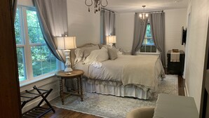 A bedroom with a bed, bedside lamps, a wooden side table, a chair, a window with curtains, and a chandelier. - Renovated Cottage - Walk to Downtown Mentone (Mentone)