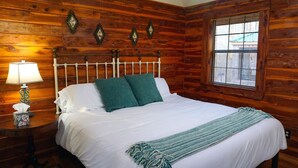 A wooden cabin-style bedroom with a bed, a nightstand with a lamp, and a window with blinds. - Couples Getaway Near Hot Springs-Jacuzzi and Hot Tub Overlooking Mountains! (Bismarck)