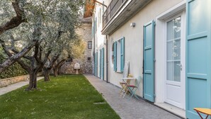 A courtyard with a grassy lawn, a stone wall, and a building with blue shutters and a white door. - Apartment "Ontano" in Agriturismo Style with Wi-Fi, Garden, and Shared Pool (Desenzano)