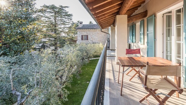 Ein Balkon mit Holztisch und Stühlen, ein Steinhaus im Hintergrund und ein Garten mit verschiedenen Pflanzen.