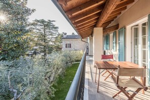 Un balcón con una mesa y sillas de madera, un edificio de piedra al fondo y un jardín con diversas plantas.