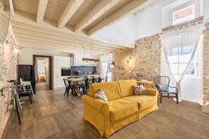 A living room with a yellow sofa, a dining area with a table and chairs, a kitchen in the background, and a stone wall.