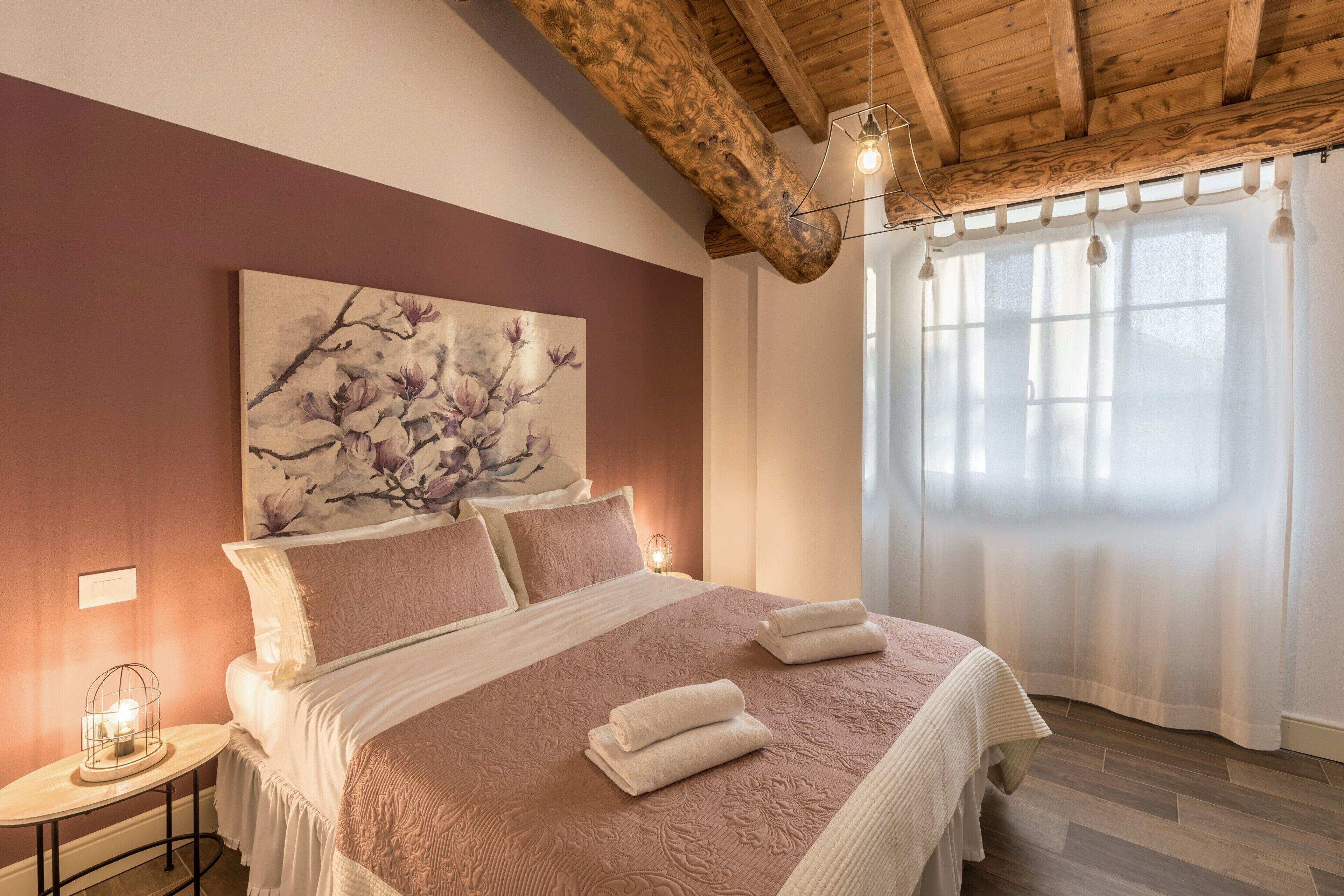 A bedroom with a large bed, a nightstand, a painting, and a window with curtains.