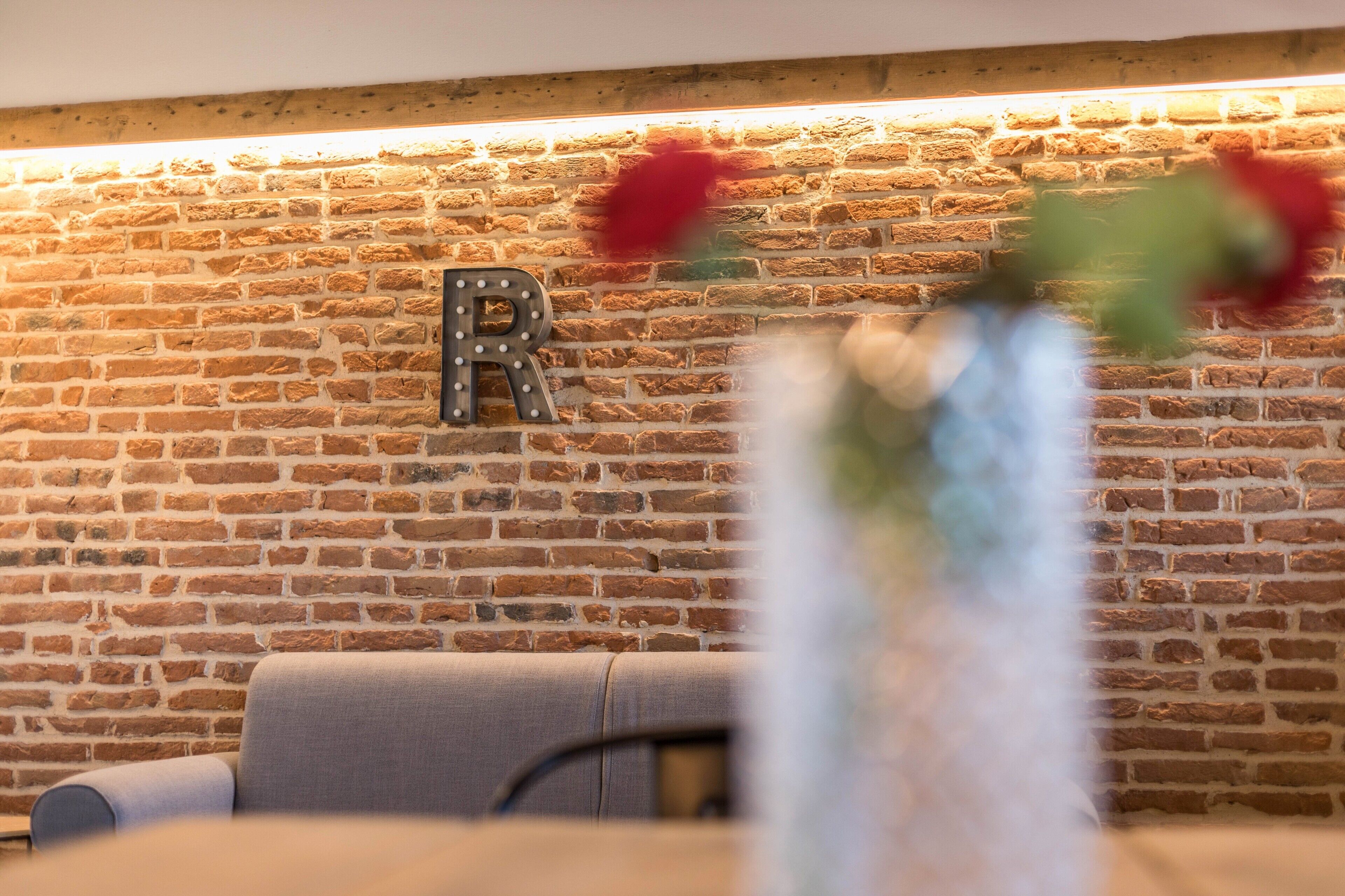 Un mur de briques sur lequel est fixée une lettre « R », un canapé gris et un vase avec des fleurs au premier plan.